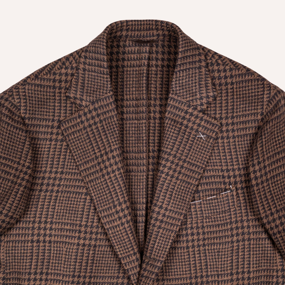 A close-up of a brown and black houndstooth patterned blazer, showing the textured fabric, collar, and lapels against a light off-white background.