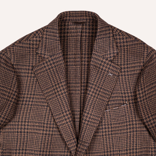 A close-up of a brown and black houndstooth patterned blazer, showing the textured fabric, collar, and lapels against a light off-white background.