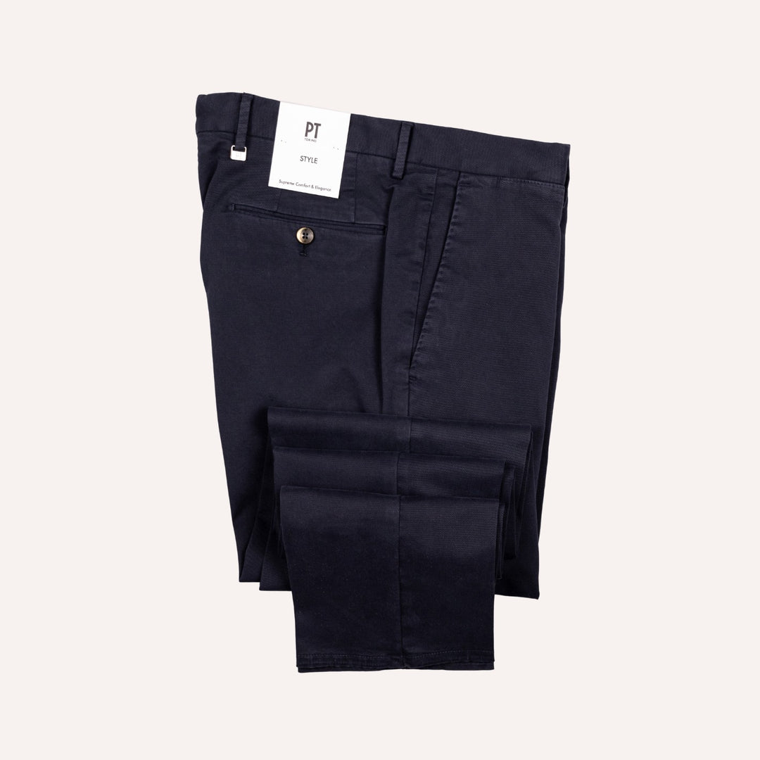 A pair of dark navy blue chino pants folded neatly, with a white brand tag reading 'PT' and model 'Style' attached near the waistband, set against a solid off-white background.