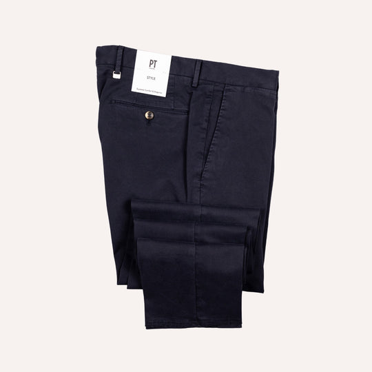 A pair of dark navy blue chino pants folded neatly, with a white brand tag reading 'PT' and model 'Style' attached near the waistband, set against a solid off-white background.