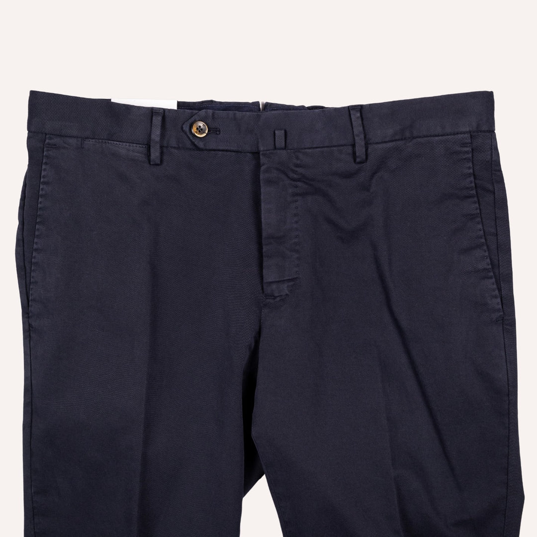 A close-up of dark navy blue trousers showing the waistband, belt loops, button closure, and front pocket on a light off-white background.