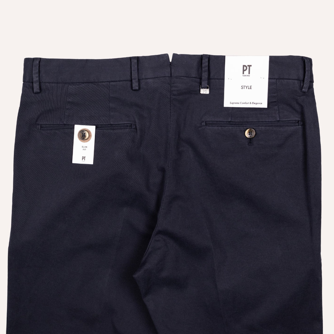 Back view of a pair of dark navy blue trousers with belt loops, back pockets, and white PT brand tags attached, set against a light beige background.