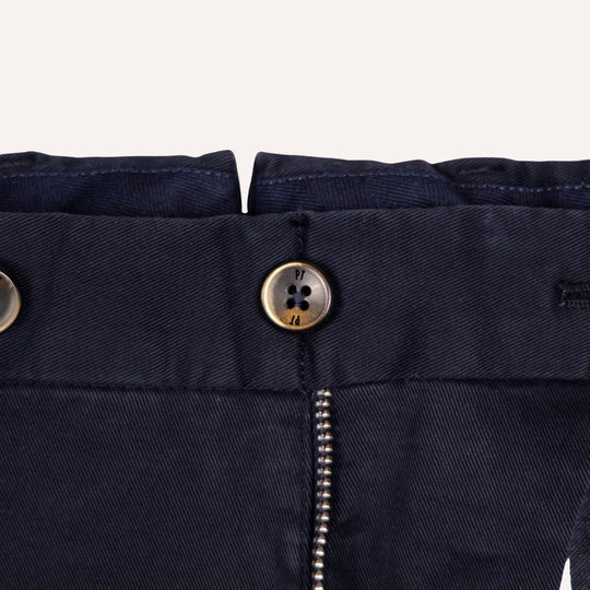 Close-up of navy blue trousers showing the waistband, a metal button closure, and a silver zipper, on a light off-white background.