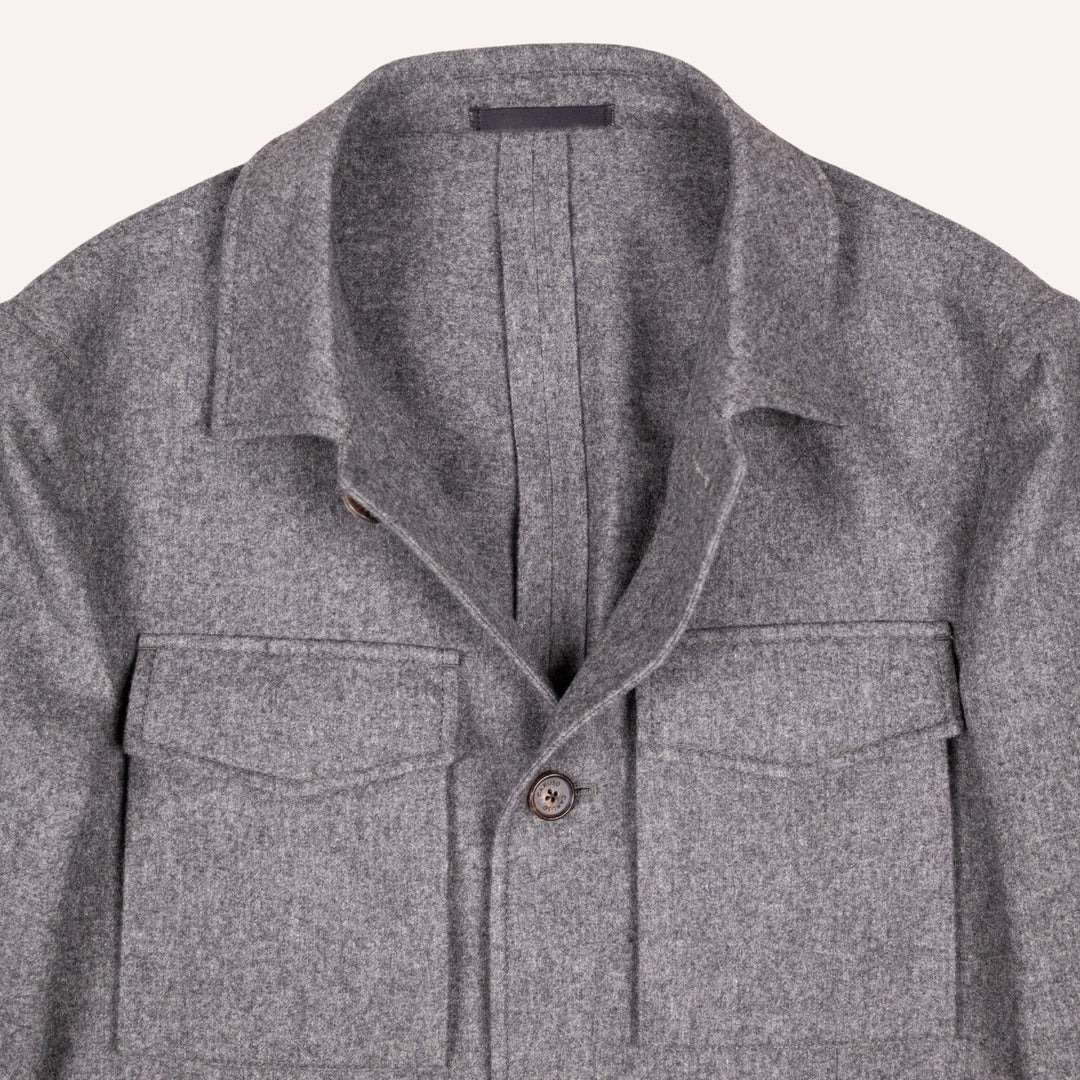 Close-up of a gray textured wool overshirt jacket with a spread collar, button front, and two chest flap pockets, shown against a light off-white background.
