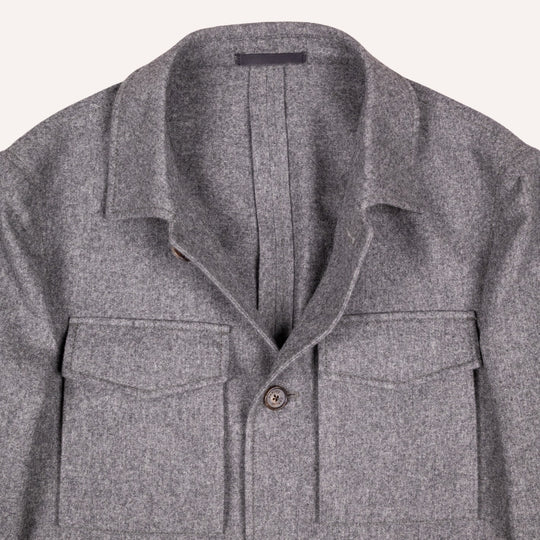 Close-up of a gray textured wool overshirt jacket with a spread collar, button front, and two chest flap pockets, shown against a light off-white background.