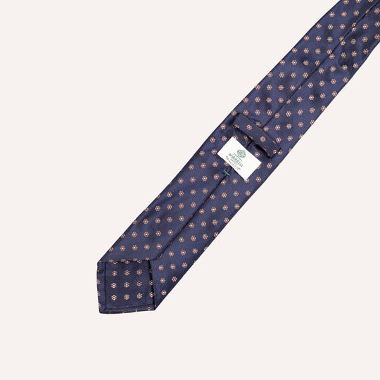 A navy blue necktie with a repeating small geometric pattern in beige and muted taupe, laid diagonally on a light cream background. A white brand label is visible near the collar area.