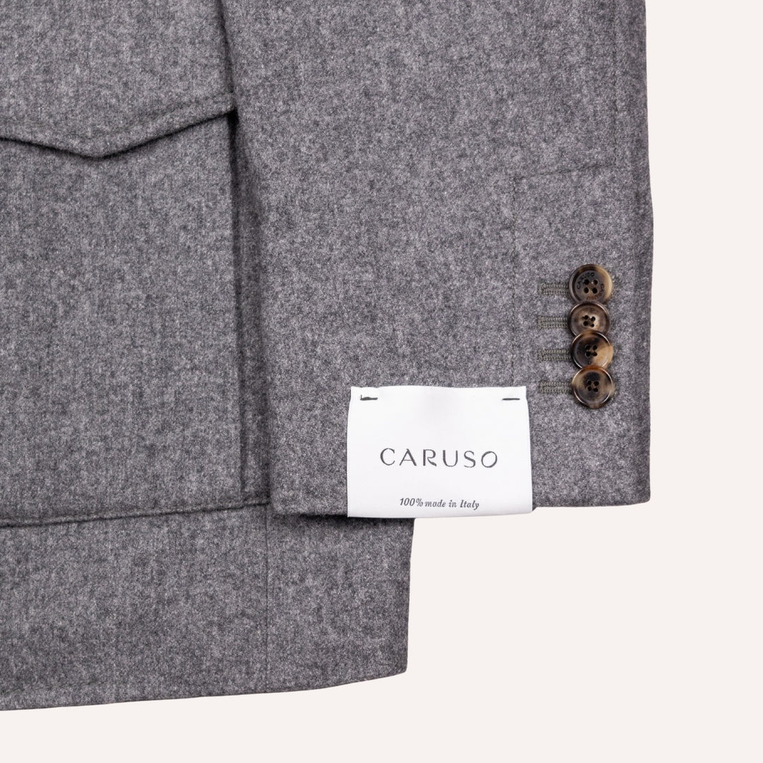 Close-up of a gray wool-blend blazer sleeve with three brown buttons and a white CARUSO brand label sewn inside the cuff, shown on a light beige background.