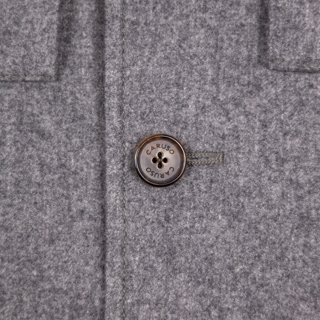 Close-up of a gray textured wool or felt garment with a round metallic button at the center. The button is engraved with the brand name 'CARUSO´
