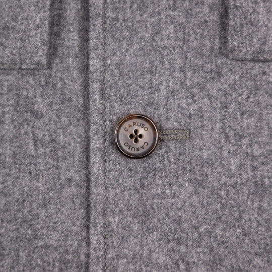 Close-up of a gray textured wool or felt garment with a round metallic button at the center. The button is engraved with the brand name 'CARUSO´