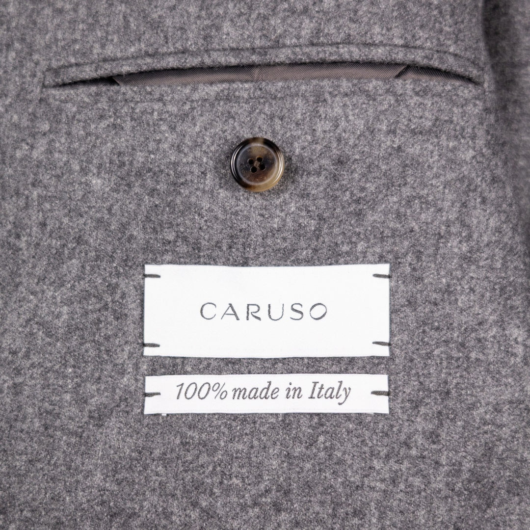 Close-up of a gray wool-blend jacket fabric showing a button, a white sewn label reading CARUSO, and another white label reading 100% made in Italy.