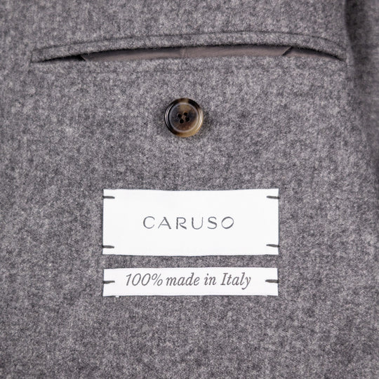 Close-up of a gray wool-blend jacket fabric showing a button, a white sewn label reading CARUSO, and another white label reading 100% made in Italy.