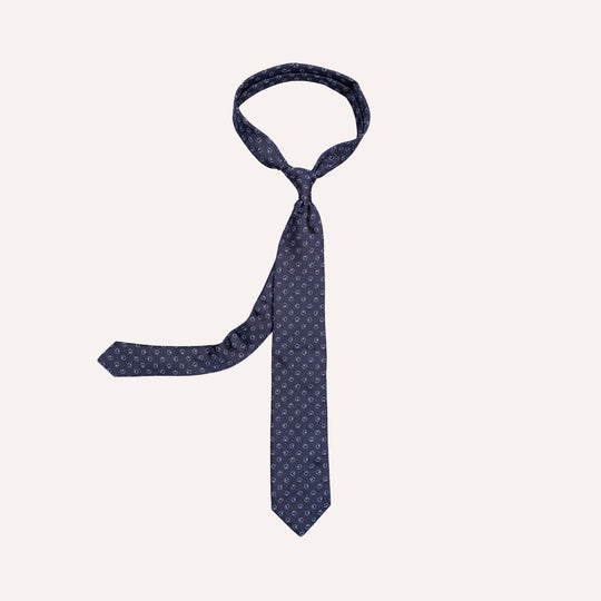 A navy blue necktie with a small repeating white geometric pattern, neatly knotted and displayed against a solid off-white background.