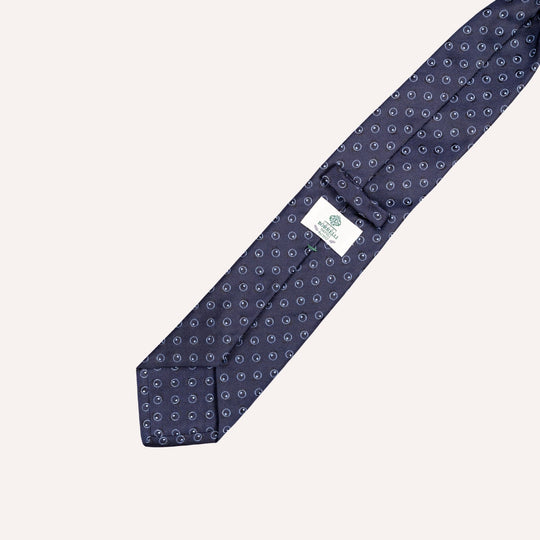 A navy blue silk necktie with a repeating small white and light blue circular pattern, laid diagonally on a light cream background. A white fabric brand tag is visible near the tip.