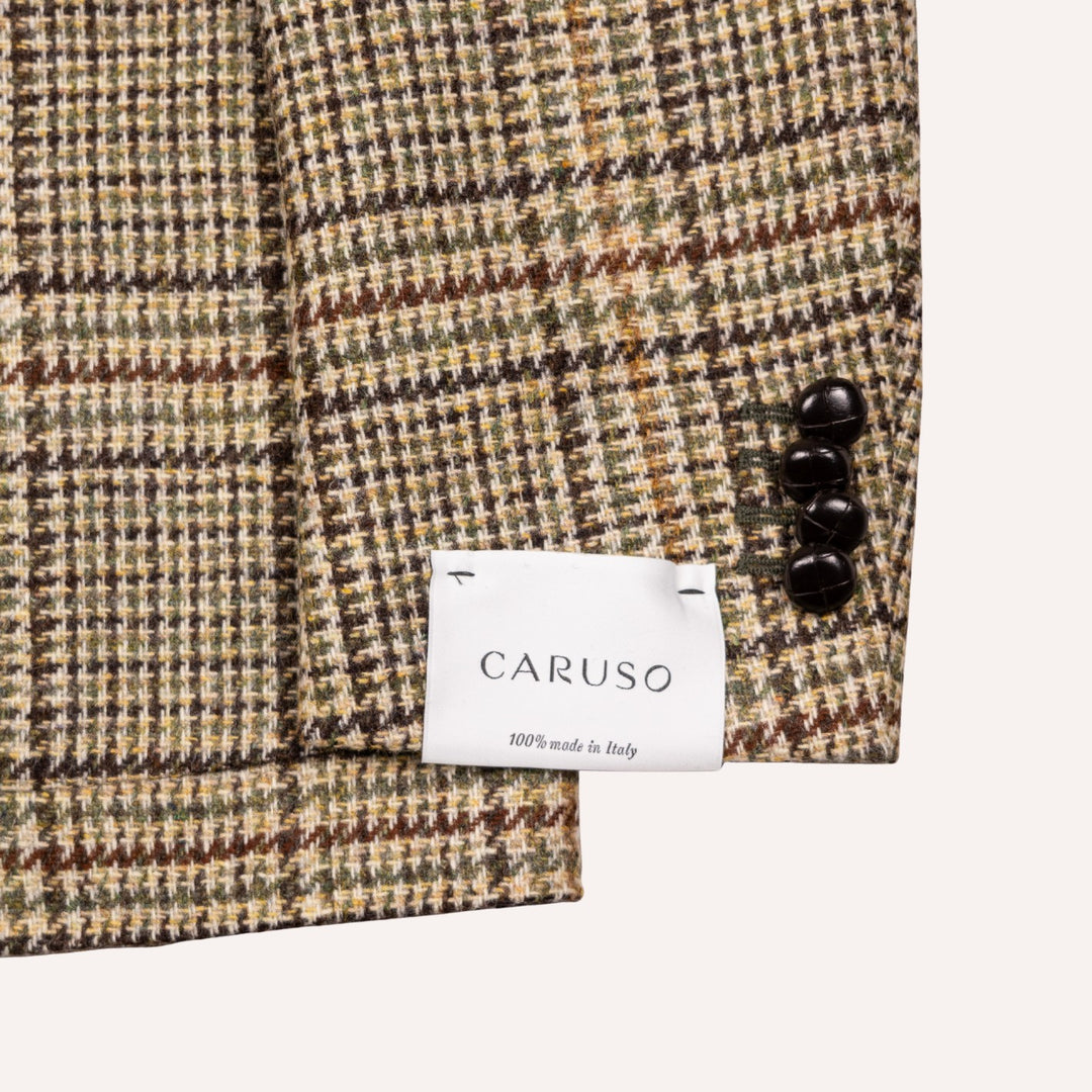 Close-up of a Caruso woven fabric with a subtle houndstooth pattern in beige, brown, and olive green, featuring three dark buttons and a white brand label.