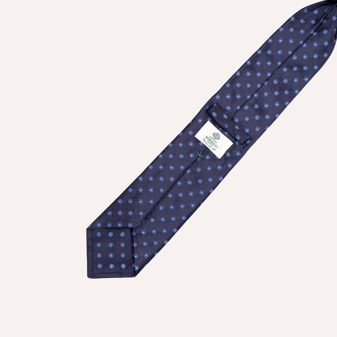 A navy blue necktie with a small light blue geometric pattern, laid flat on a light beige background. A white brand label is visible near the collar area.