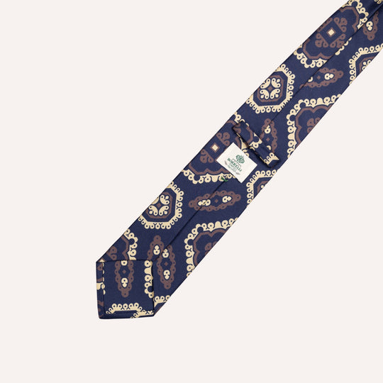 A navy blue silk necktie with a repeating white and light blue circular pattern, laid diagonally on a light cream background. A white brand tag is visible near the collar end.