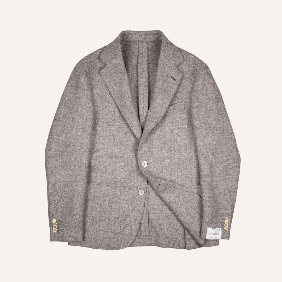 A light gray textured tweed blazer with notched lapels, two front buttons, and cream tone button accents on the cuffs, laid flat against a light cream background.