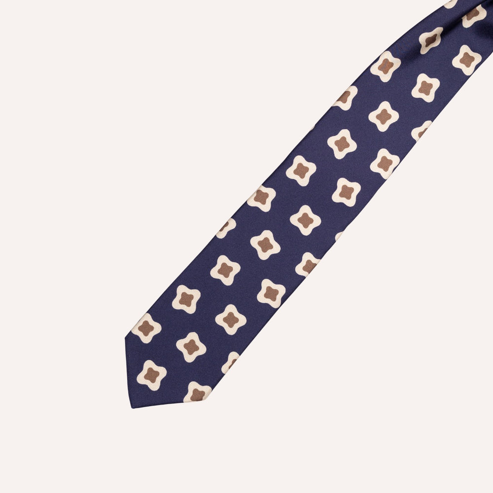 A navy blue necktie with a repeating geometric pattern of cream and brown four-pointed starburst motifs, laid diagonally on a light beige background.