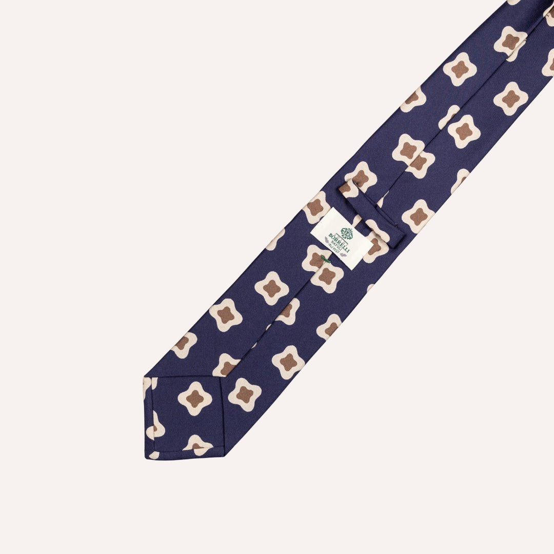 A navy blue necktie with a repeating geometric quatrefoil pattern in beige and brown, displayed diagonally on a light cream background.