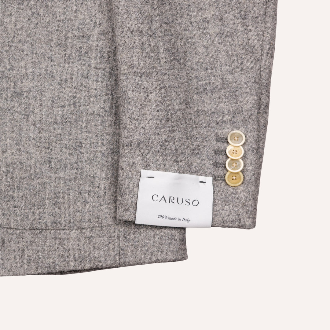 Close-up of a gray wool-blend blazer sleeve showing the cuff, three light wood buttons, and a white CARUSO brand label sewn onto the inside of the cuff.