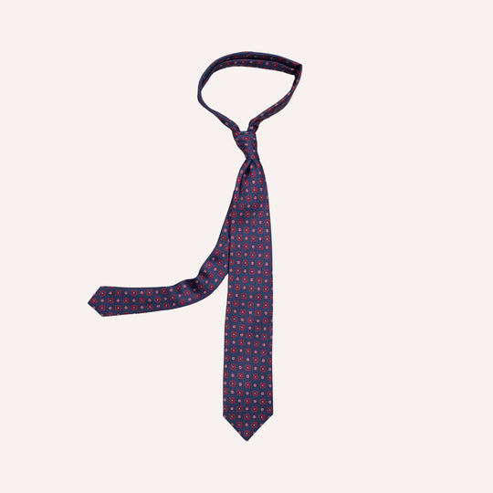 A navy blue necktie with a small repeating medallion pattern of red and white dots, tied in a standard knot and displayed against a solid off-white background.