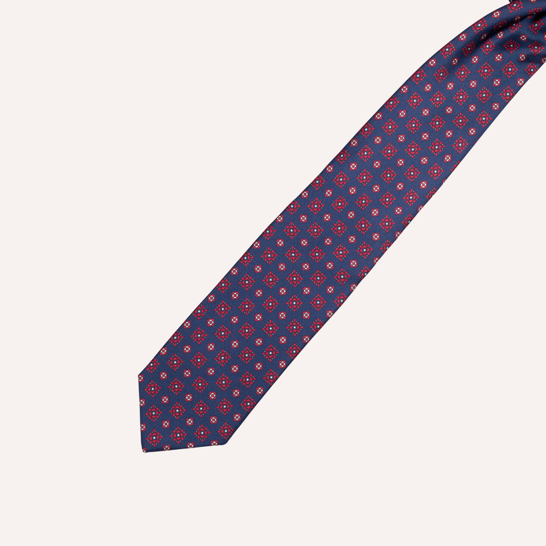 A navy blue necktie with a repeating medallion red and white geometric pattern, laid diagonally on a light cream background.