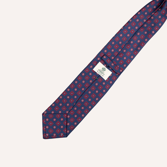 A navy blue silk necktie with a repeating medallion white and light blue circular pattern, laid diagonally on a light cream background. A white brand tag is visible near the collar end.