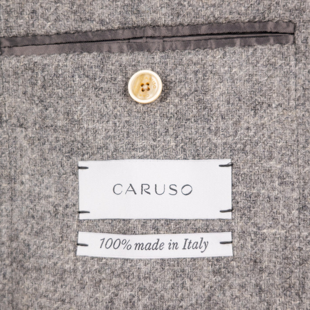 Close-up of a gray textured fabric surface with a white CARUSO label and a second label reading '100% made in Italy', plus a small cream button above.