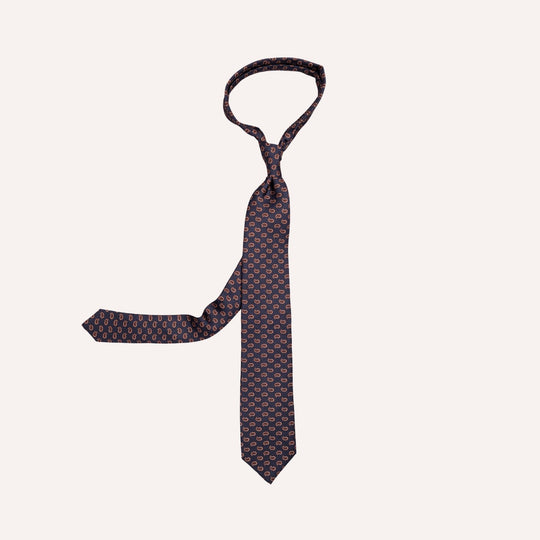 A dark navy blue necktie with a repeating small taupe and brown geometric paisley pattern, displayed flat against a light cream background.