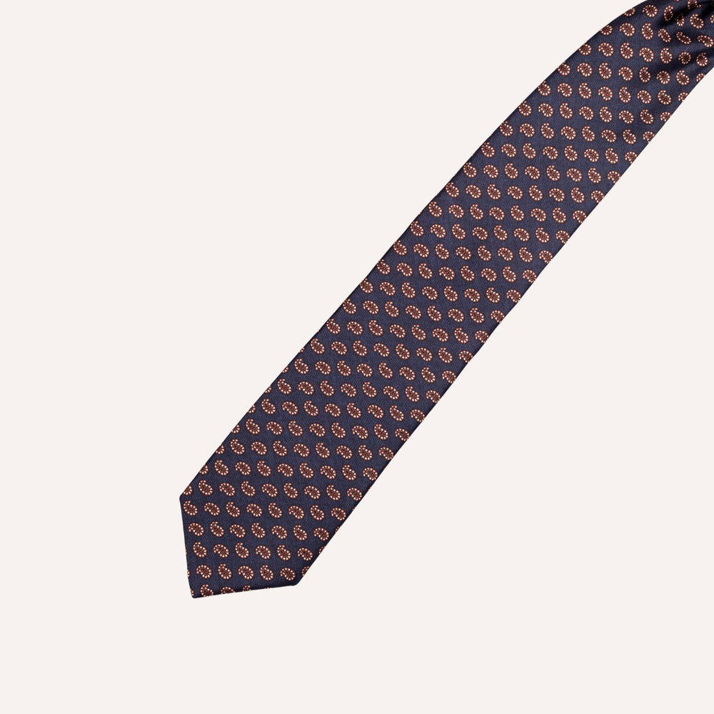 A classic necktie with a repeating paisley pattern in brown and white on a dark navy blue background, laid out diagonally on a light cream background.