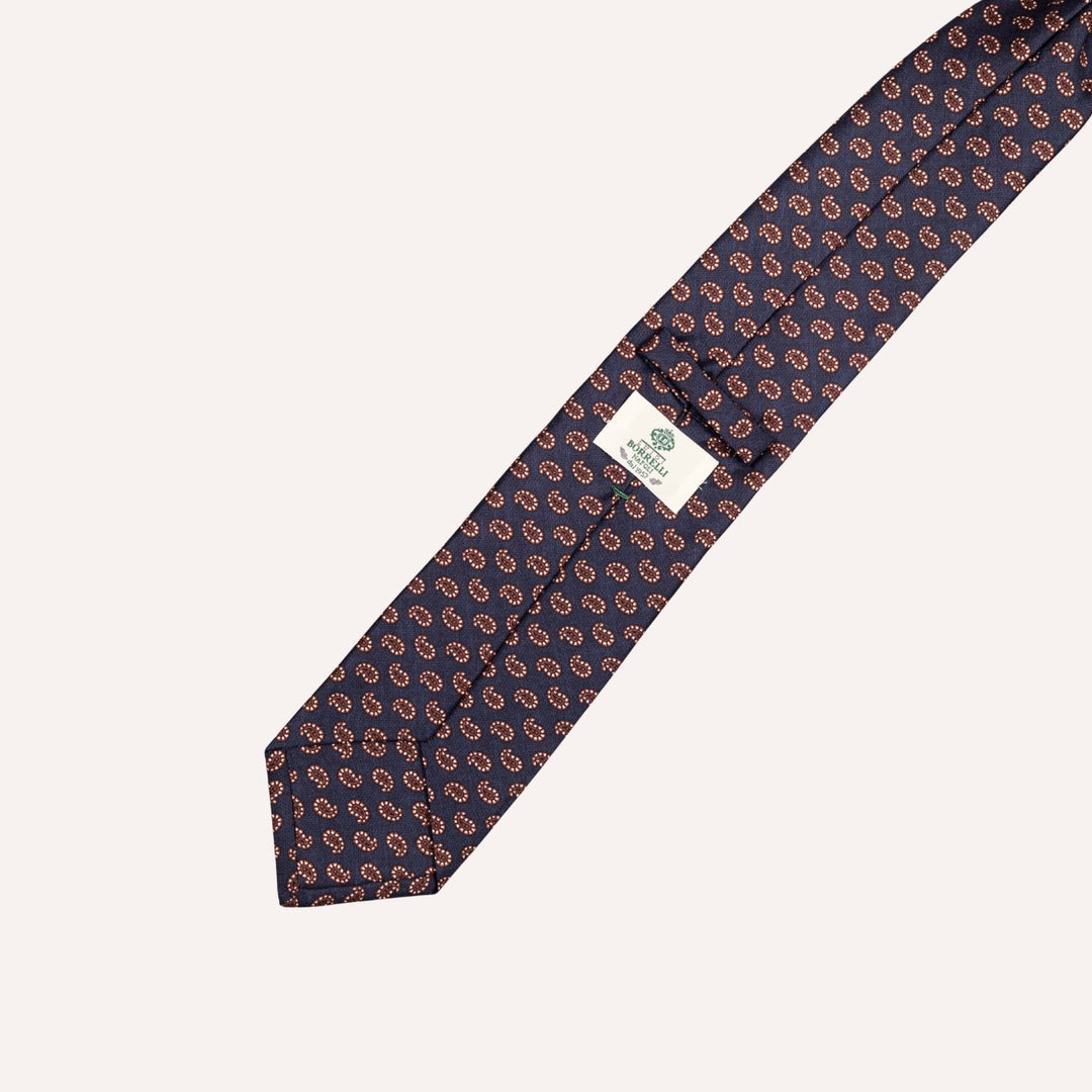 A navy blue silk necktie with a repeating paisley pattern in brown and white, featuring a white brand label with green text.