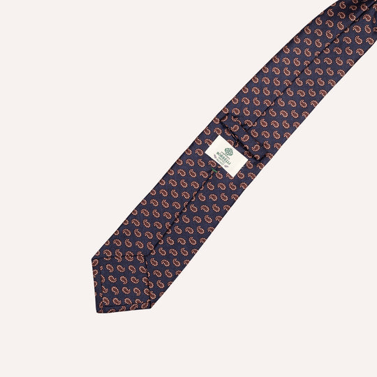 A navy blue silk necktie with a repeating paisley pattern in brown and white, featuring a white brand label with green text.