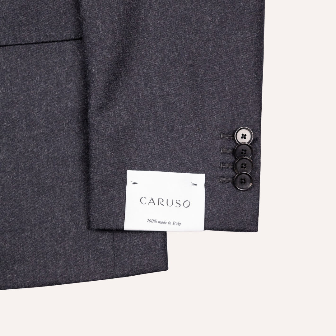 Close-up of a dark gray wool suit jacket showing the fabric texture, a white brand label reading CARUSO, and three buttons on the sleeve cuff against a light cream background.