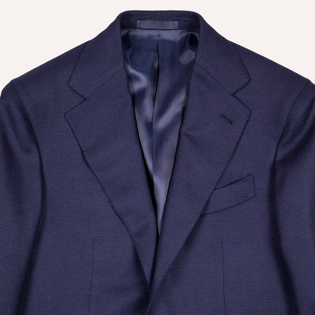 A close-up of a navy blue textured suit jacket, showing the lapels, outer pocket, and a satin lining in a matching dark blue color, set against a plain off-white background.