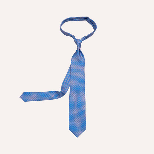 A blue necktie with a small geometric pattern, laid flat on a light cream background.