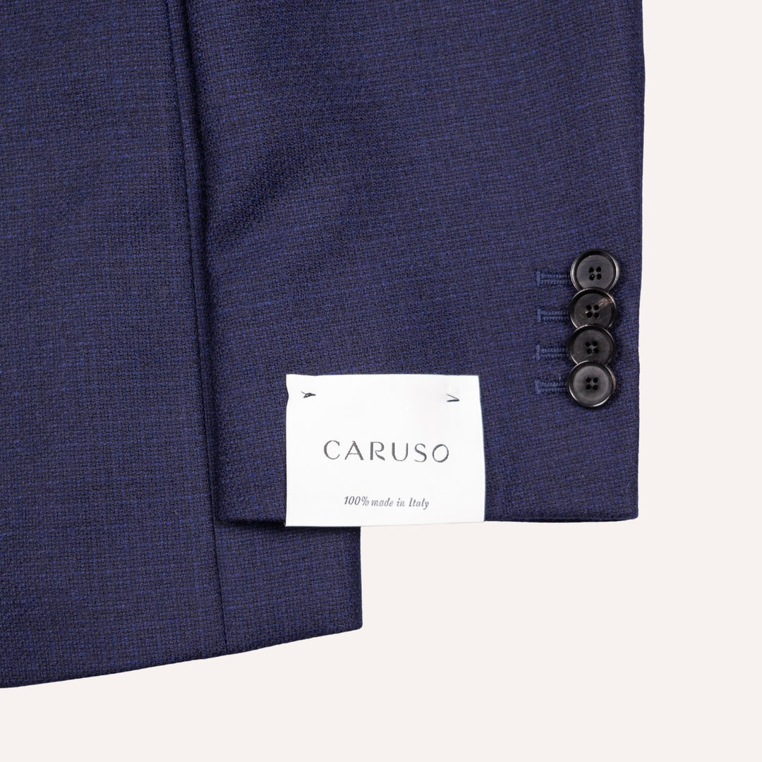 Close-up of a navy blue textured suit jacket sleeve showing three buttons on the cuff and a white brand label reading CARUSO.