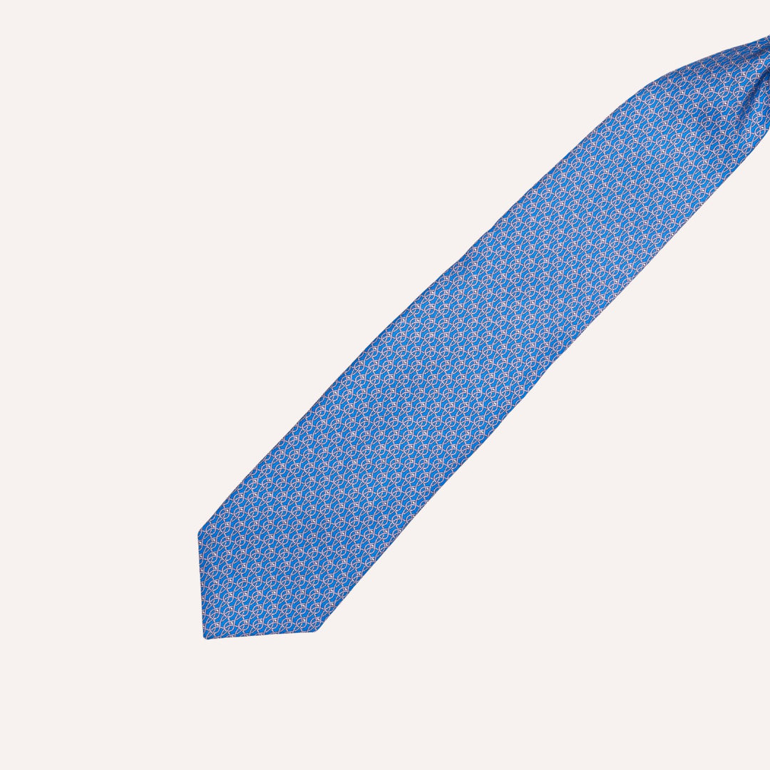A blue necktie with a repeating small geometric pattern in white and light blue, laid diagonally on a plain off-white background.