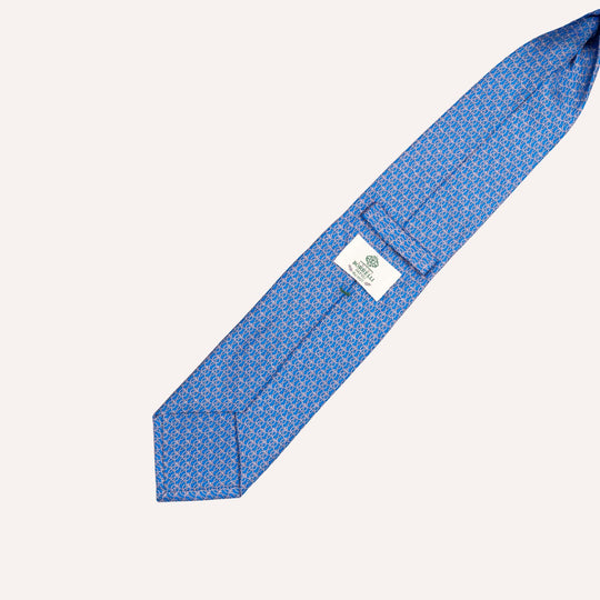 A blue silk necktie with a repeating small white and light blue geometric pattern, laid diagonally on a light cream background. A white brand label is visible near the center.