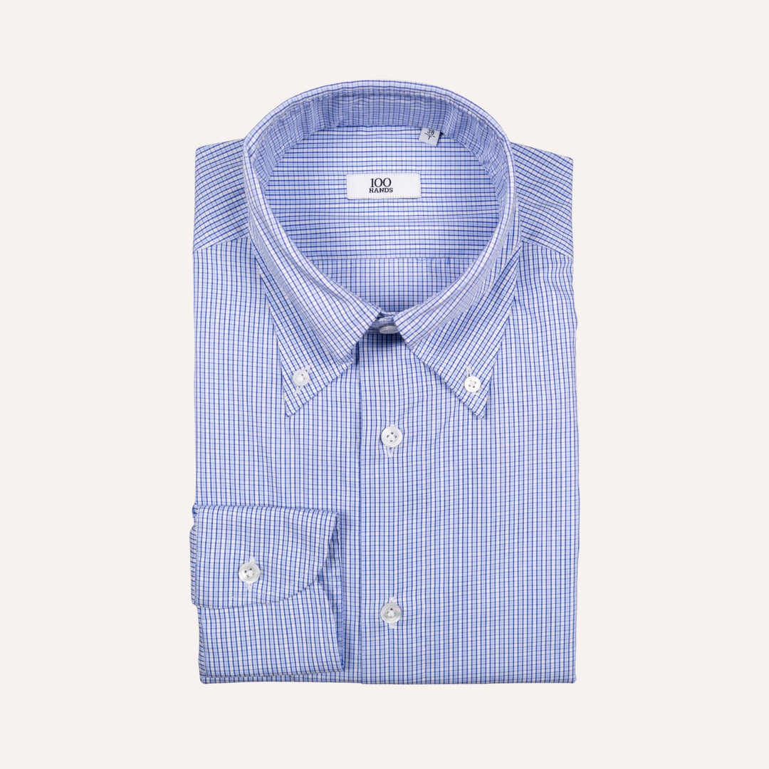 A folded blue and white gingham button-down shirt with a point collar and a single cuff, displayed on a light cream background.