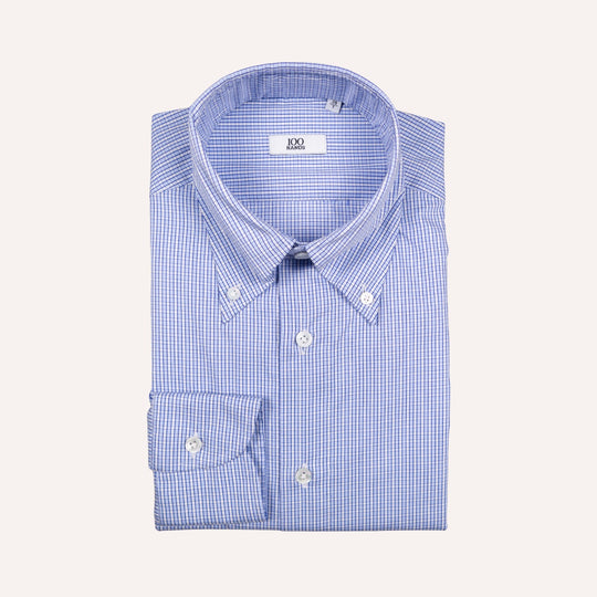 A folded blue and white gingham button-down shirt with a point collar and a single cuff, displayed on a light cream background.