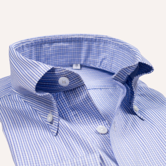 Close-up of a blue and white micro-check patterned button-down shirt collar with white buttons, set against a clean off-white background.