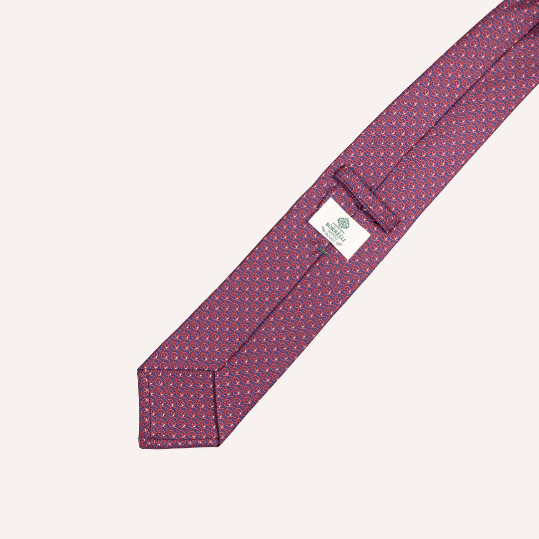 A close-up of a necktie with a purple and red micro-pattern, laid diagonally on a light cream background. A white brand label for 'The Necktie Company' is visible near the tip.