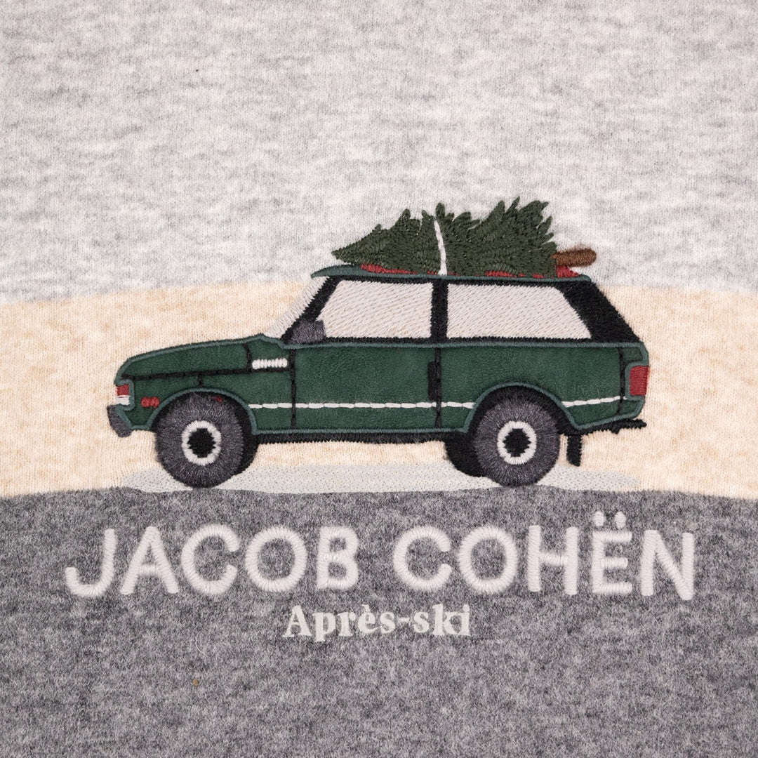 A close-up of a heather gray fabric surface featuring an embroidered design of a dark green Range Rover carrying a Christmas tree on its roof, with the text 'JACOB COHÉN' and 'Après-ski' below. 