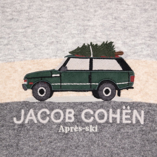 A close-up of a heather gray fabric surface featuring an embroidered design of a dark green Range Rover carrying a Christmas tree on its roof, with the text 'JACOB COHÉN' and 'Après-ski' below. 