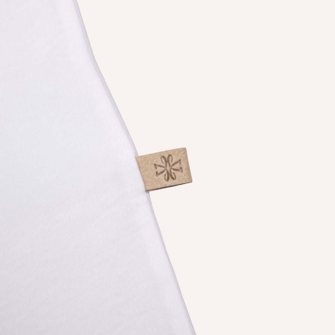 A close-up of a white garment corner with a small beige fabric tag attached, featuring a debossed floral or snowflake-like symbol.