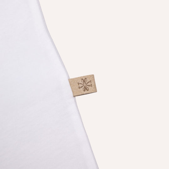 A close-up of a white garment corner with a small beige fabric tag attached, featuring a debossed floral or snowflake-like symbol.