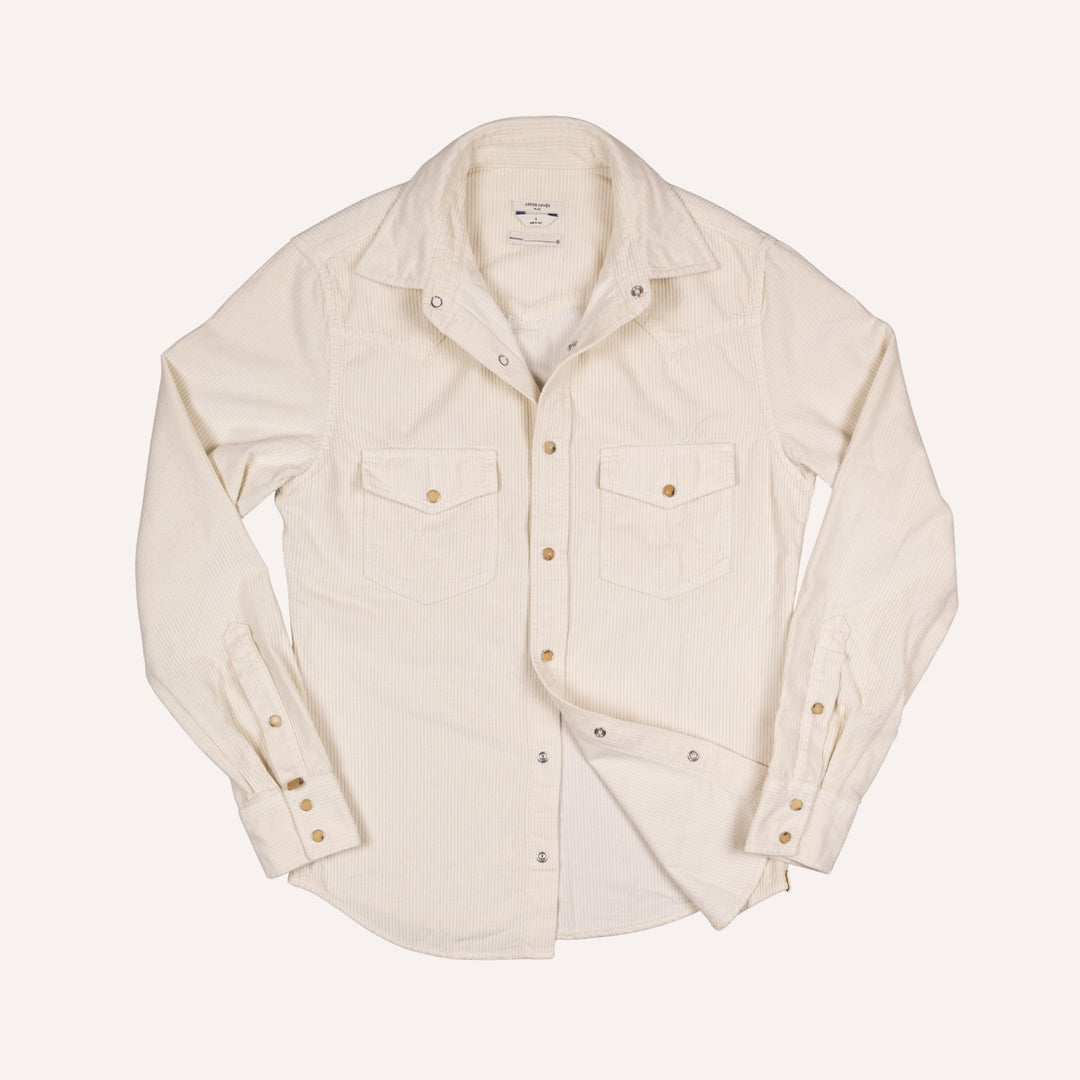 An off-white long-sleeve corduroy western shirt with two chest flap pockets and brass buttons, laid flat against a light cream background.
