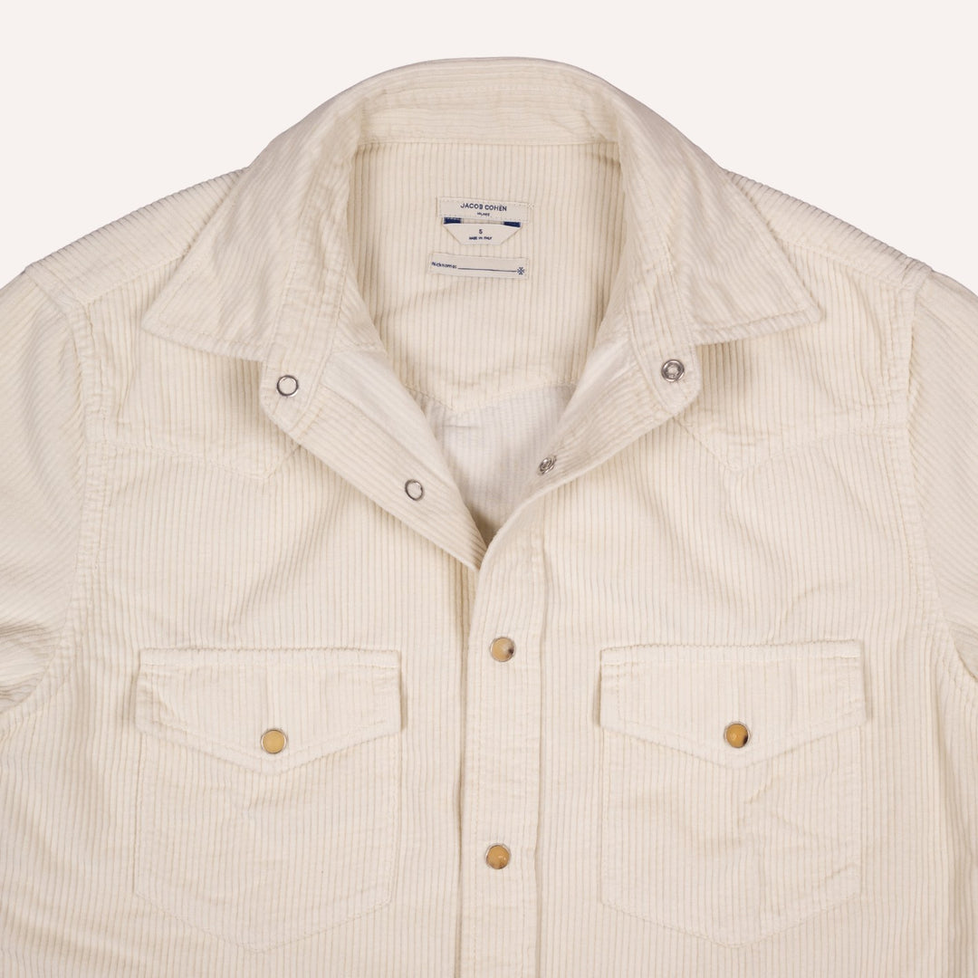 A close-up of a cream-colored corduroy shirt jacket with two buttoned chest pockets and a pointed collar, shown against a light off-white background.