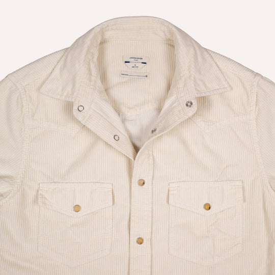 A close-up of a cream-colored corduroy shirt jacket with two buttoned chest pockets and a pointed collar, shown against a light off-white background.