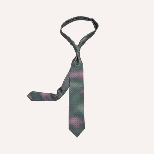 A slim necktie with a fine micro-pattern of small white dots on a dark slate gray-green background, presented in a loose knot on a light cream background.